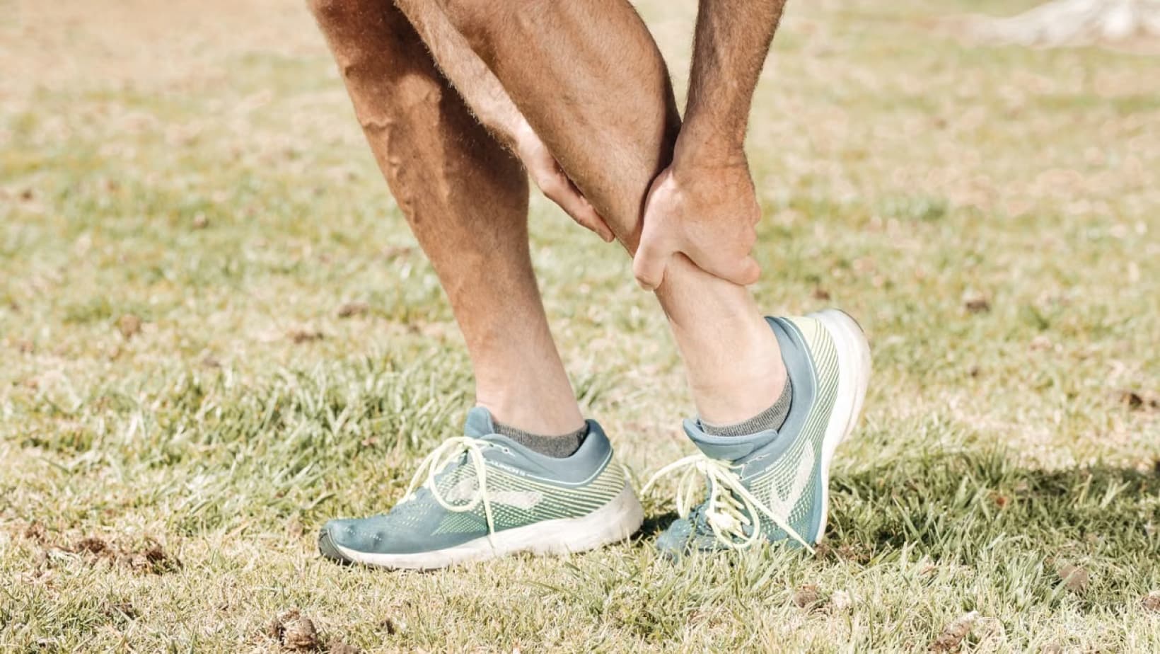 Runner in sports shoes holding a sore lower leg on the grass